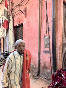 Cosa tur Marrakech. En teppehandler går rundt med et teppe over skulderen. Bakgrunnen er rosa, fra medinaen.