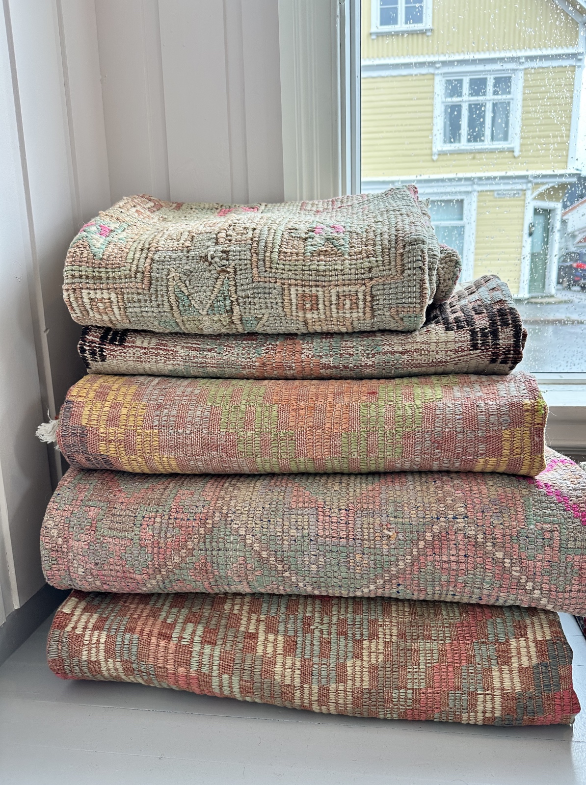 Stablede vintage kilimtepper i dempede rosa, grønne og oransje toner, fotografert ved et regnvått vindu i Cosa showroom