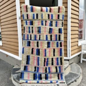 Et marokkansk berberteppe hengende foran et gult hus i trehusbebyggelsen i kristiansand. teppet er i klare farger som blå, rosa, kremhvit, sort og hvit.
