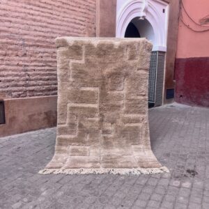 Håndknyttet Beni Ourain-teppe i beige ull med geometrisk skulpturelt mønster. Vist utendørs i en marokkansk medina med rosa murvegger. Mål 250x154 cm, pris 12000,-