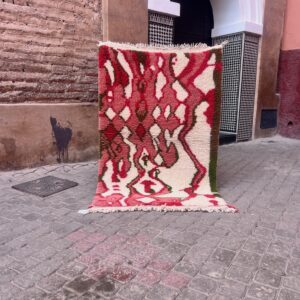 Håndknyttet marokkansk Boujaad teppe i rosa, rødt og oliven holdt opp foran en tradisjonell marokkansk dør. Mål 160x105 cm, pris 5500,-