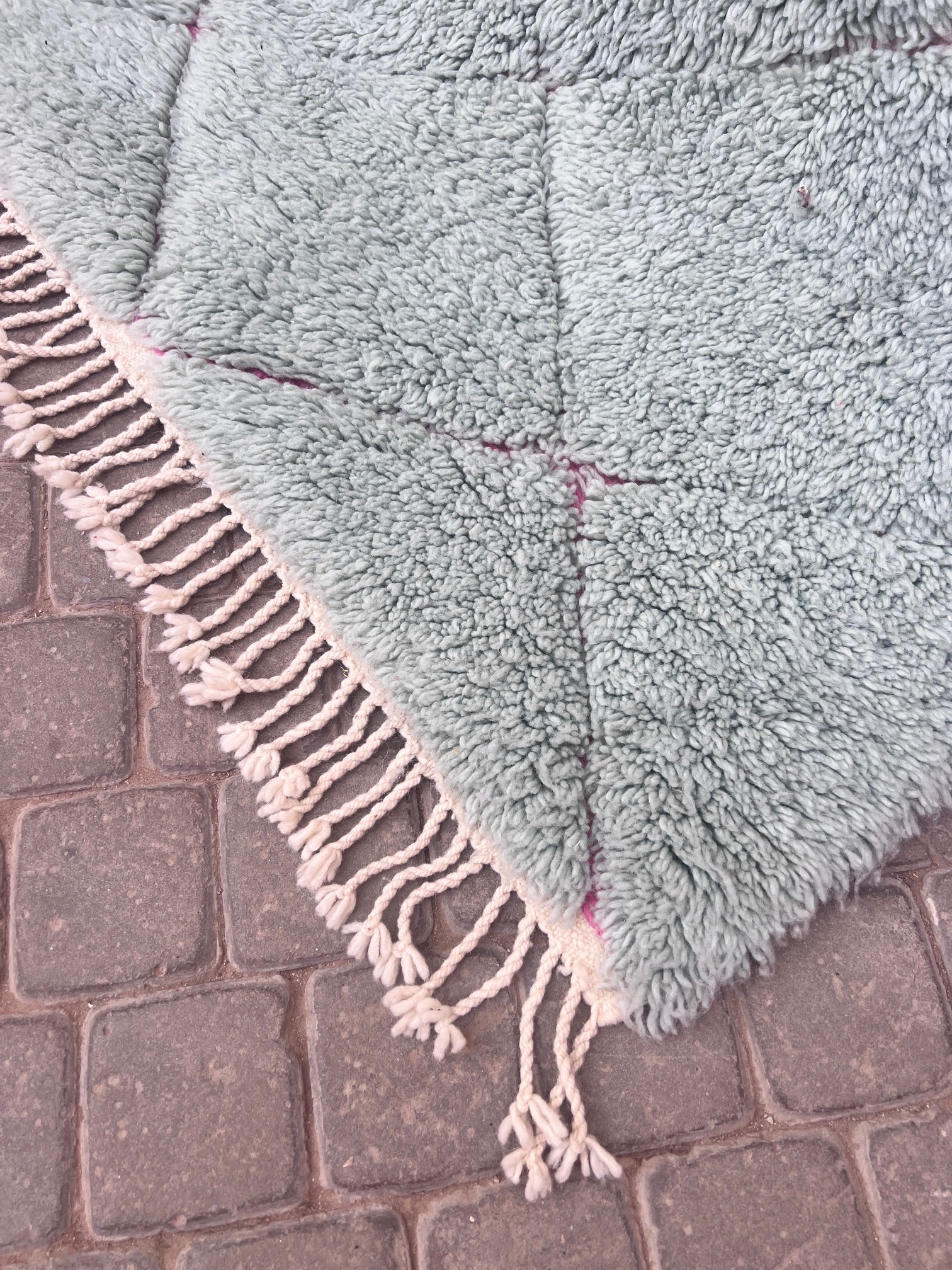 Lyseblått beni Ourain teppe med rosa diamantmønster og hvite frynser, holdt opp foran en port i Marrakech. Mål 157x108 cm, pris 5500,-