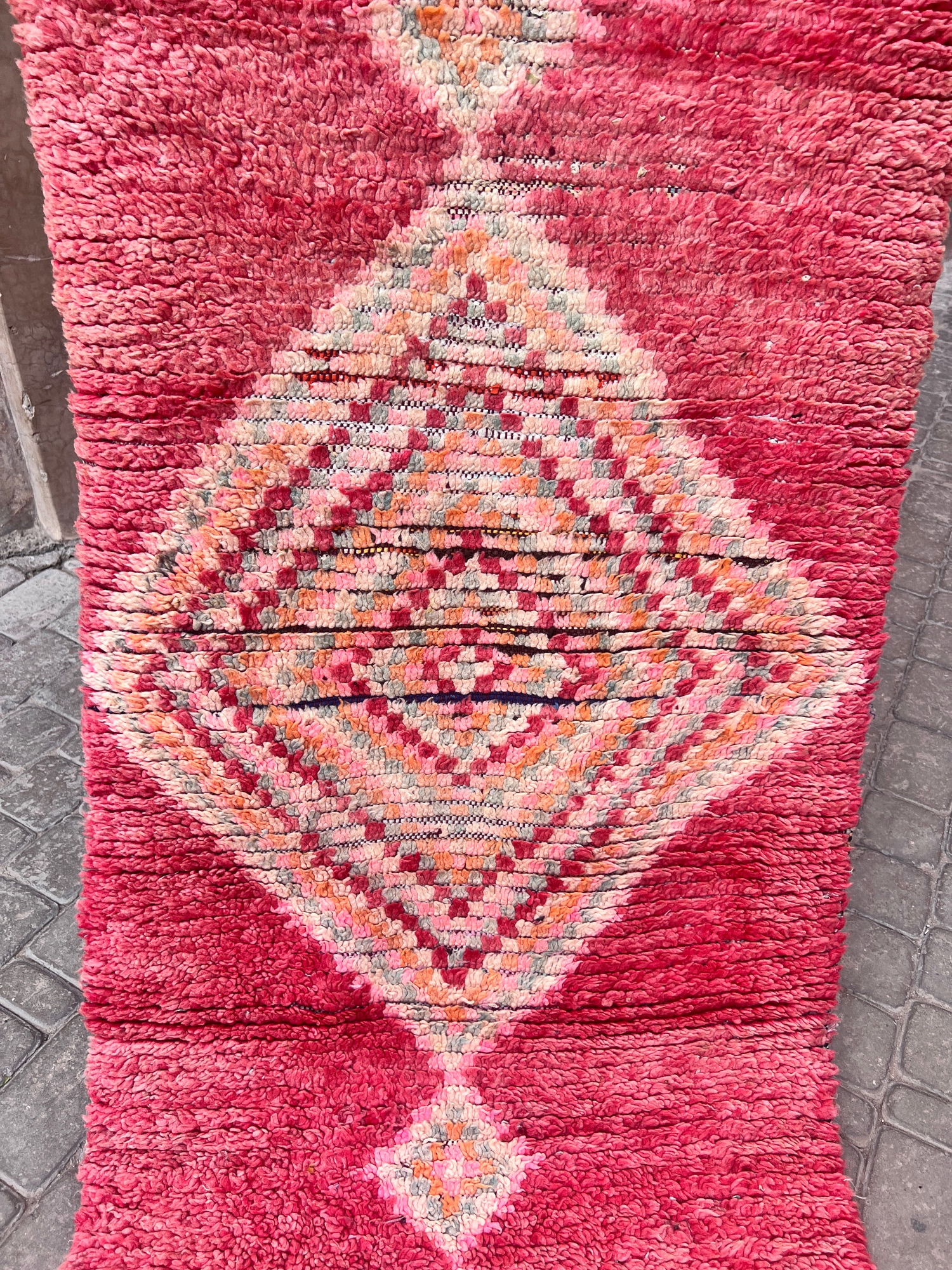 En håndknyttet vintage-løper fra Boujaad regionen i Marokko. Rød, rosa, dus grønn og aprikos diamantmønster. Teppet holdes opp foran rosa murvegger i en gate i Marrakech