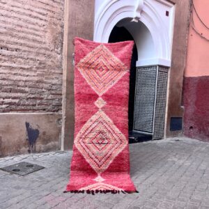 En håndknyttet vintage-løper fra Boujaad regionen i Marokko. Rød, rosa, dus grønn og aprikos diamantmønster. Teppet holdes opp foran rosa murvegger i en gate i Marrakech