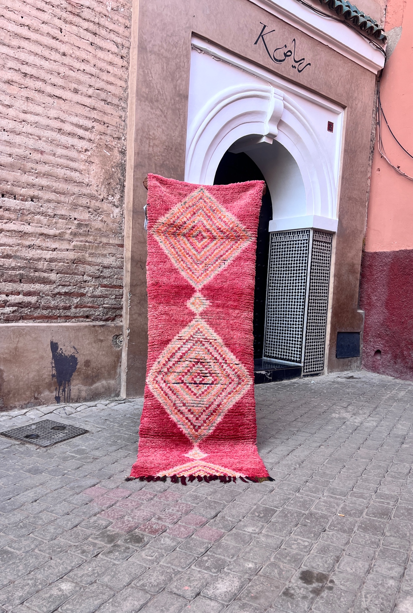 En håndknyttet vintage-løper fra Boujaad regionen i Marokko. Rød, rosa, dus grønn og aprikos diamantmønster. Teppet holdes opp foran rosa murvegger i en gate i Marrakech