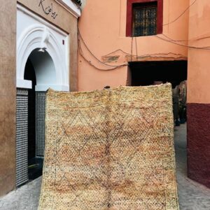 Stort vintage Beni Mguild-teppe i falmede toner av sand, aprikos og fersken, med fint rute- og diamantmønster, holdt opp i en gate i medinaen i Marrakech