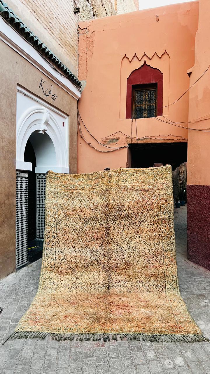 Stort vintage Beni Mguild-teppe i falmede toner av sand, aprikos og fersken, med fint rute- og diamantmønster, holdt opp i en gate i medinaen i Marrakech